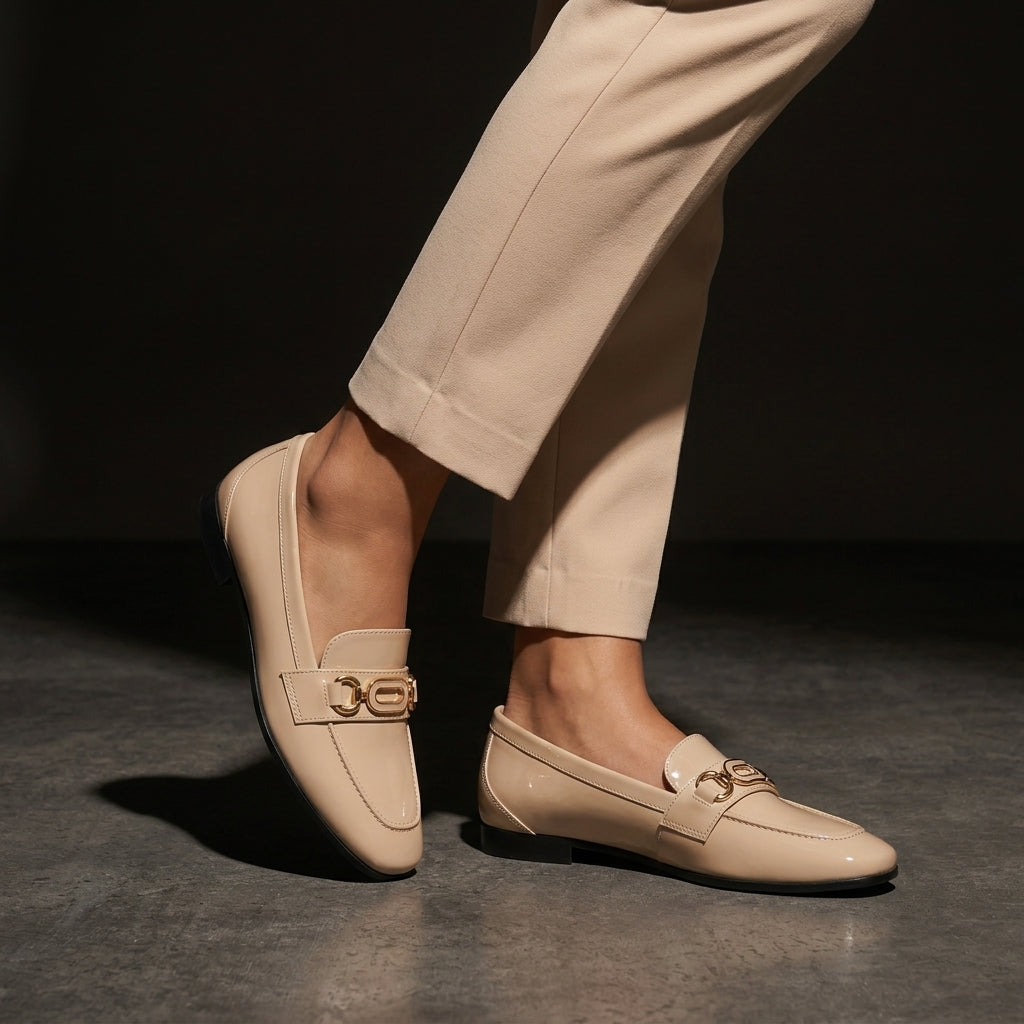 Woman wearing glossy beige patent ECADRA loafers featuring a gold-tone hardware buckle detail and matching beige trousers.