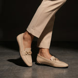 Woman wearing glossy beige patent ECADRA loafers featuring a gold-tone hardware buckle detail and matching beige trousers.
