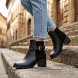 Close-up side view of classic black block heel ankle boot showing suede rear panel and inner zipper closure on stone stairs.
