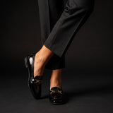Dynamic view of crossed legs modeling glossy black patent leather loafers against a dark backdrop.