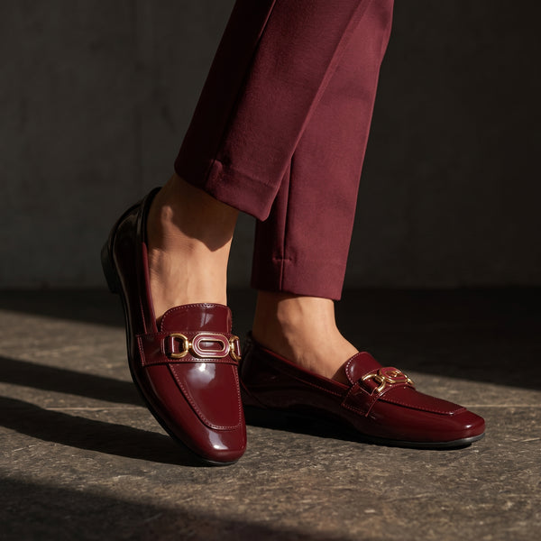 Close up side view of women's burgundy patent leather loafers standing on a textured stone floor in sunlight.