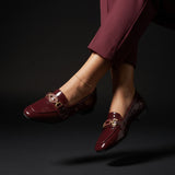 Dynamic shot of burgundy patent leather loafers with gold chain detailing worn on feet against a dark background.