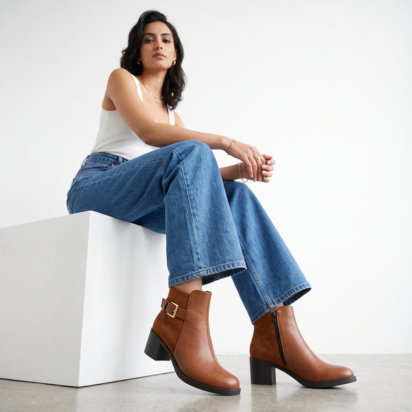 Model sitting on a white box showcasing tan brown block heel ankle boots with gold buckle and inner side zipper.