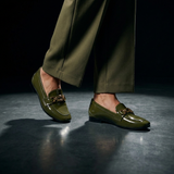 A pair of glossy olive green loafers with gold-tone hardware, shown on a model wearing matching olive trousers against a dark studio background. Olive Chain loafer Studio Shoot 2 | ECADRA