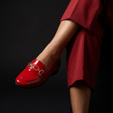 Woman seated with crossed legs wearing glossy red patent ECADRA loafers and matching red trousers.