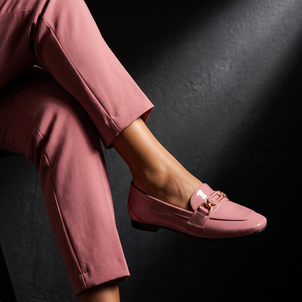 Woman sitting gracefully while wearing glossy rose patent ECADRA loafers with gold hardware buckle details and matching rose attire.