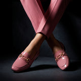 Woman seated with crossed legs wearing glossy rose patent ECADRA loafers and matching rose trousers.