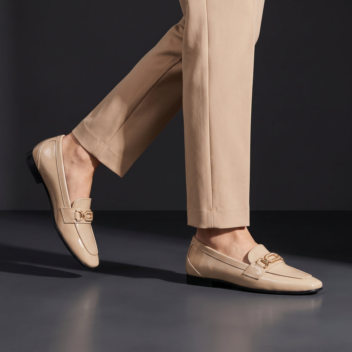 Woman wearing glossy beige patent ECADRA loafers featuring a gold-tone hardware buckle detail and matching beige trousers.