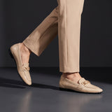 Woman wearing glossy beige patent ECADRA loafers featuring a gold-tone hardware buckle detail and matching beige trousers.
