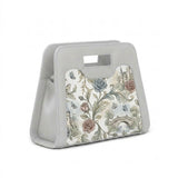A modern, grey neoprene handbag with a cutout handle, featuring a detailed vintage print of castles, roses, and butterflies, positioned next to its white shoulder strap with black clasps against a white background.