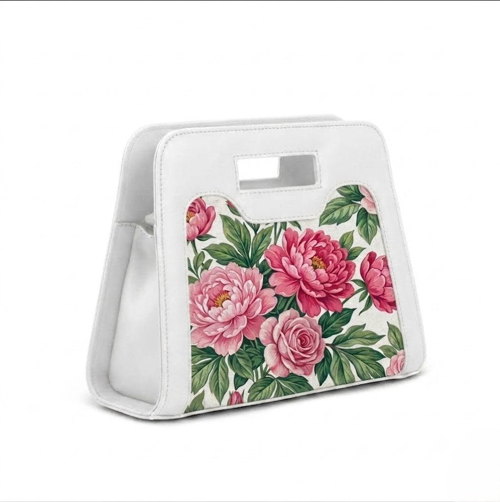 A vibrant arrangement of vintage pink peonies, roses, and green leaves is featured on the front panel of a structured white top-handle handbag with a detachable shoulder strap.
